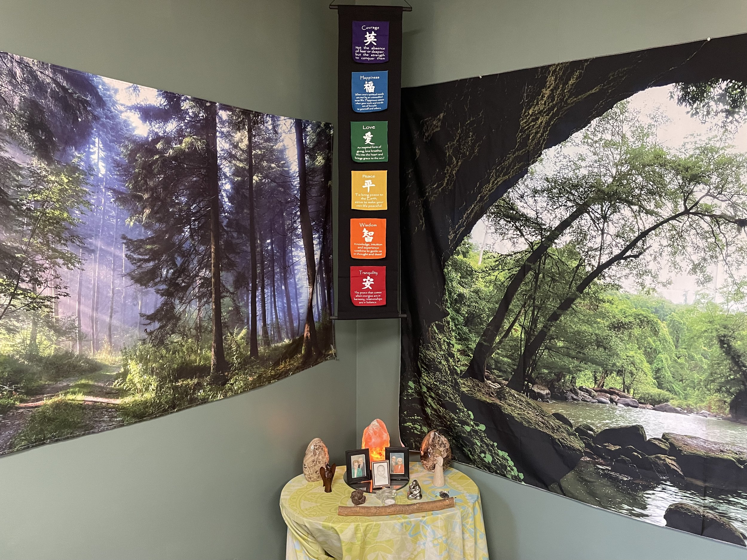 Room corner with two large nature murals and a chakra banner. Left mural depicts a sunlit forest path. Right mural shows a river lined with lush trees. Below, a small table displays crystals, a Himalayan salt lamp, and decorative items.