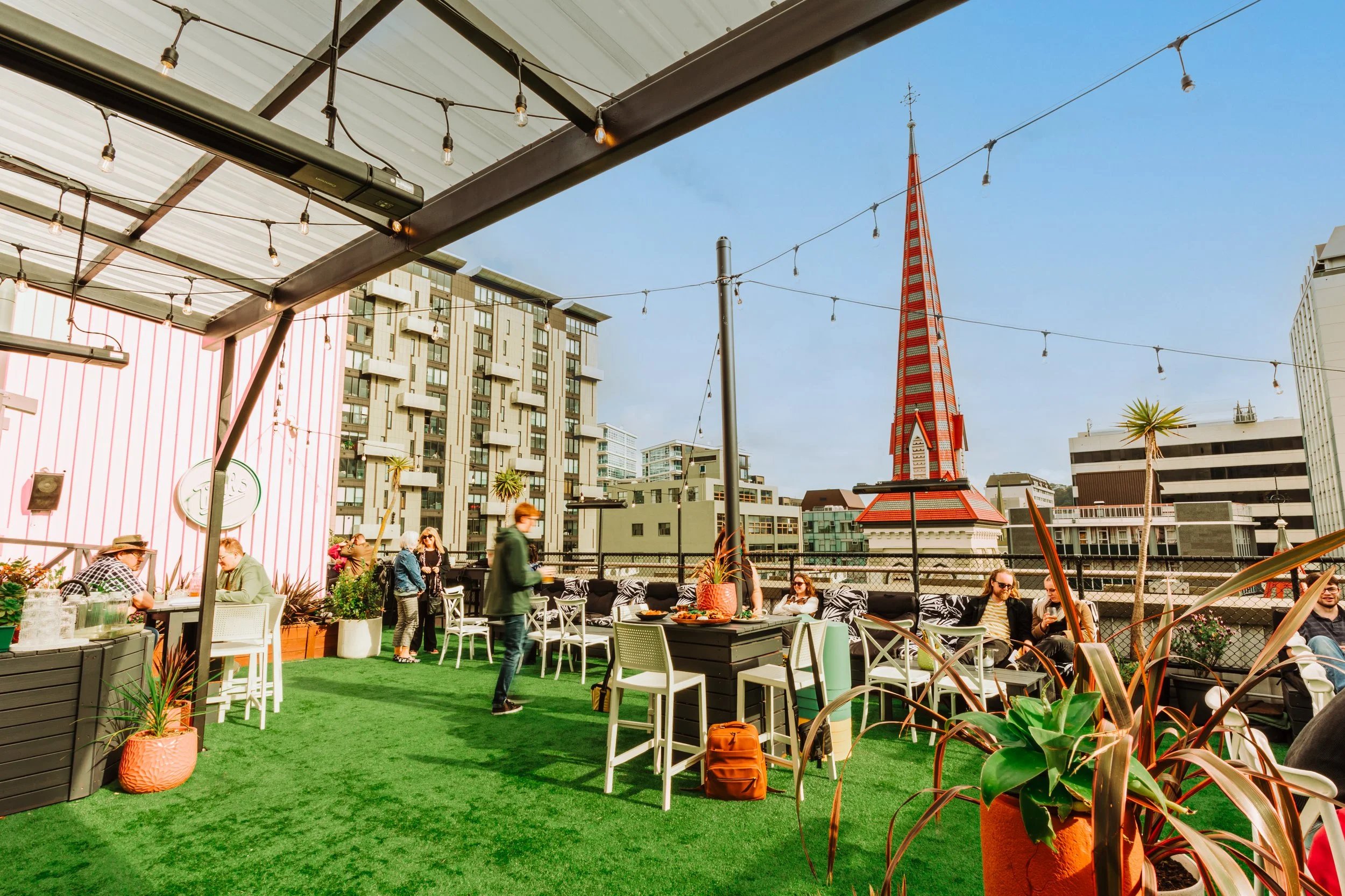 People sitting and socializing on a rooftop patio with outdoor furniture, string lights, potted plants, and city buildings in the background under a clear blue sky.
