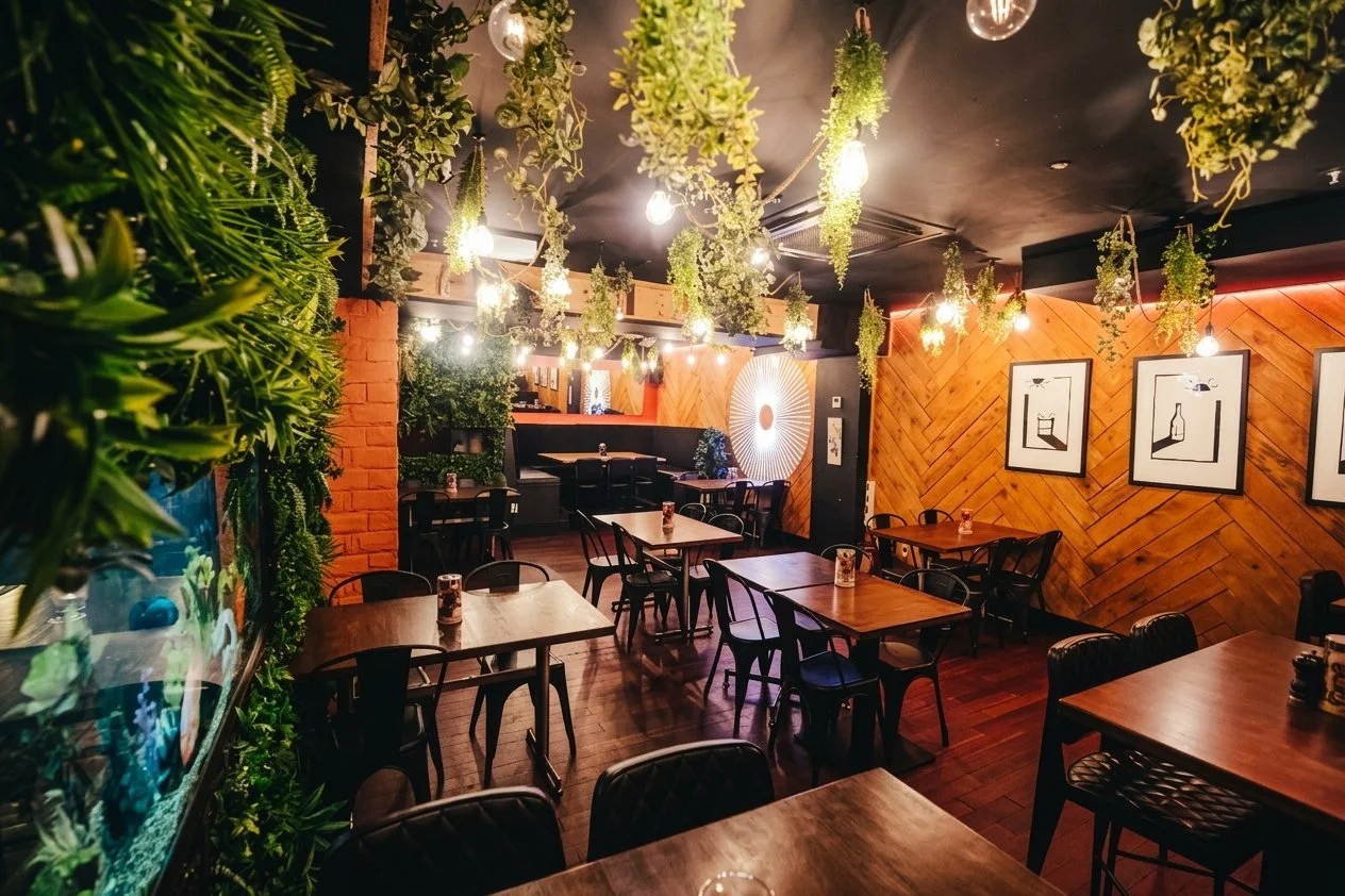 Interior of a cozy restaurant or cafe with wooden and brick walls, hanging plants, framed artwork, and dim lighting.