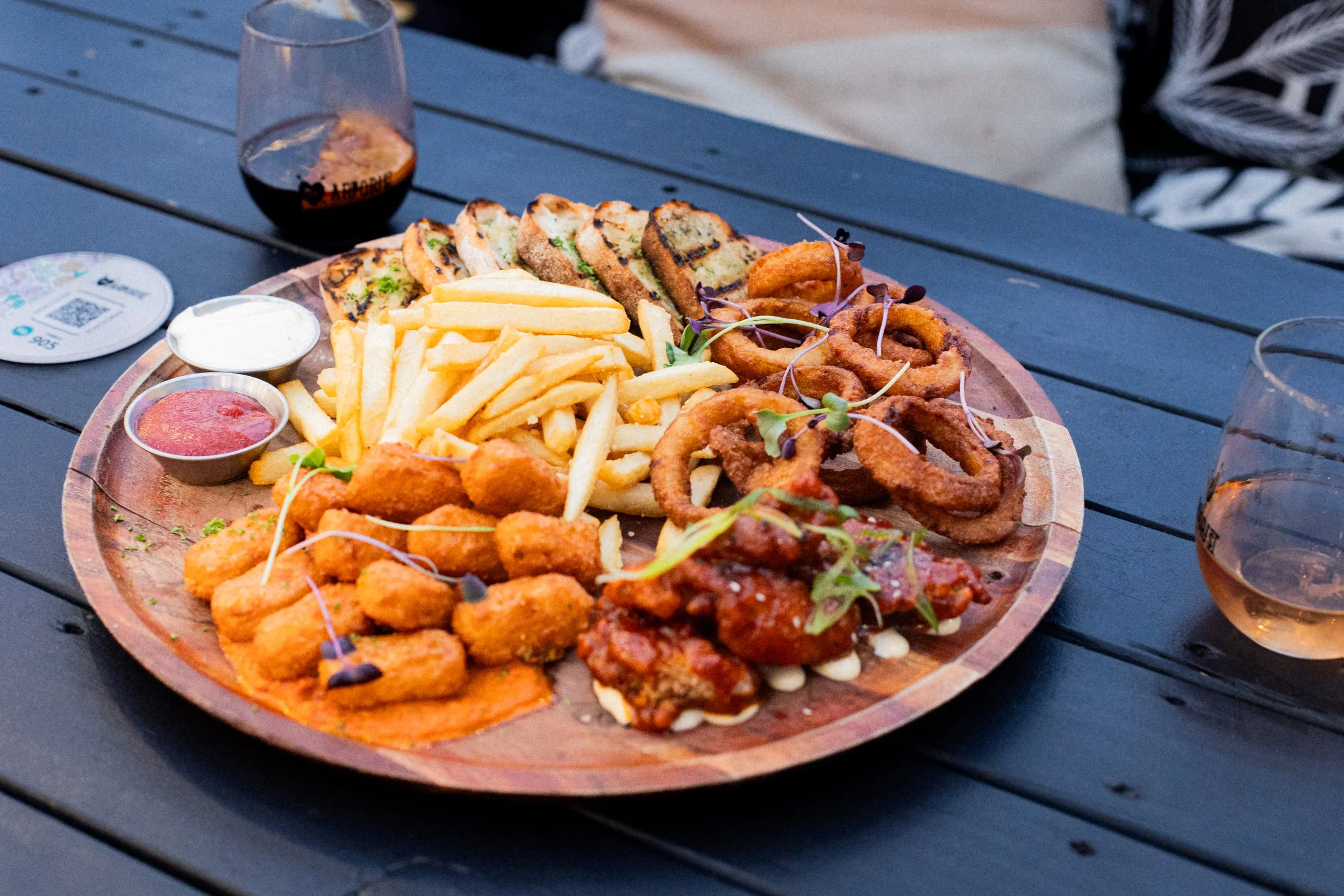 A round wooden platter with assorted fried foods, French fries, grilled bread slices, and dipping sauces, on an outdoor dark wooden table.