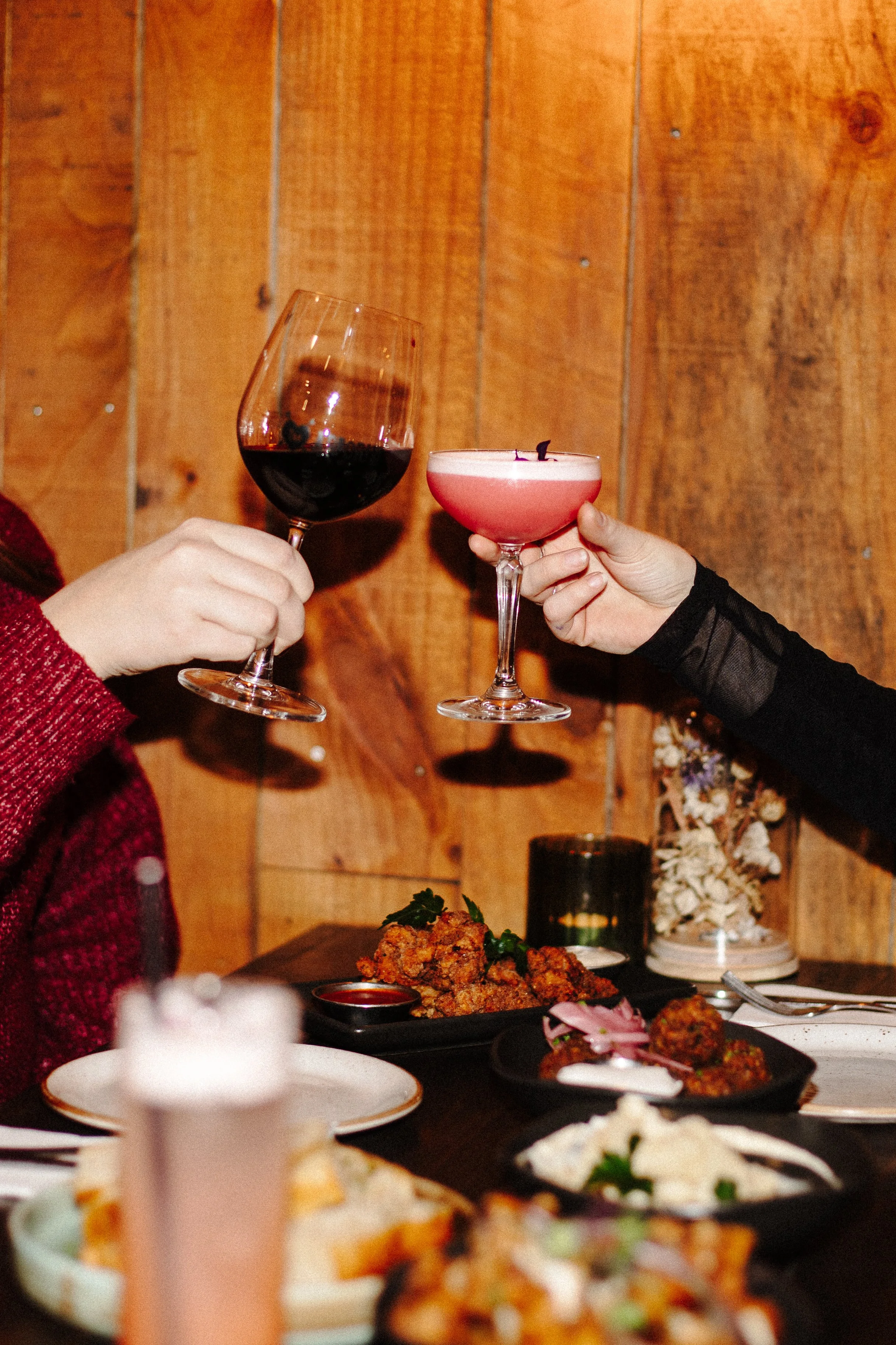 Two people toasting with glasses of red wine and pink cocktail at a dinner table with food, against wooden wall background.