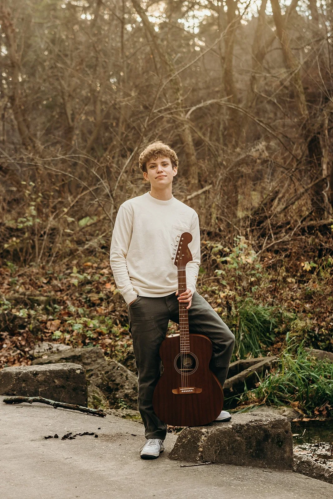 A young man with curly hair wearing a white sweatshirt and gray pants holding an acoustic guitar, standing on a rock in a wooded area during daytime.