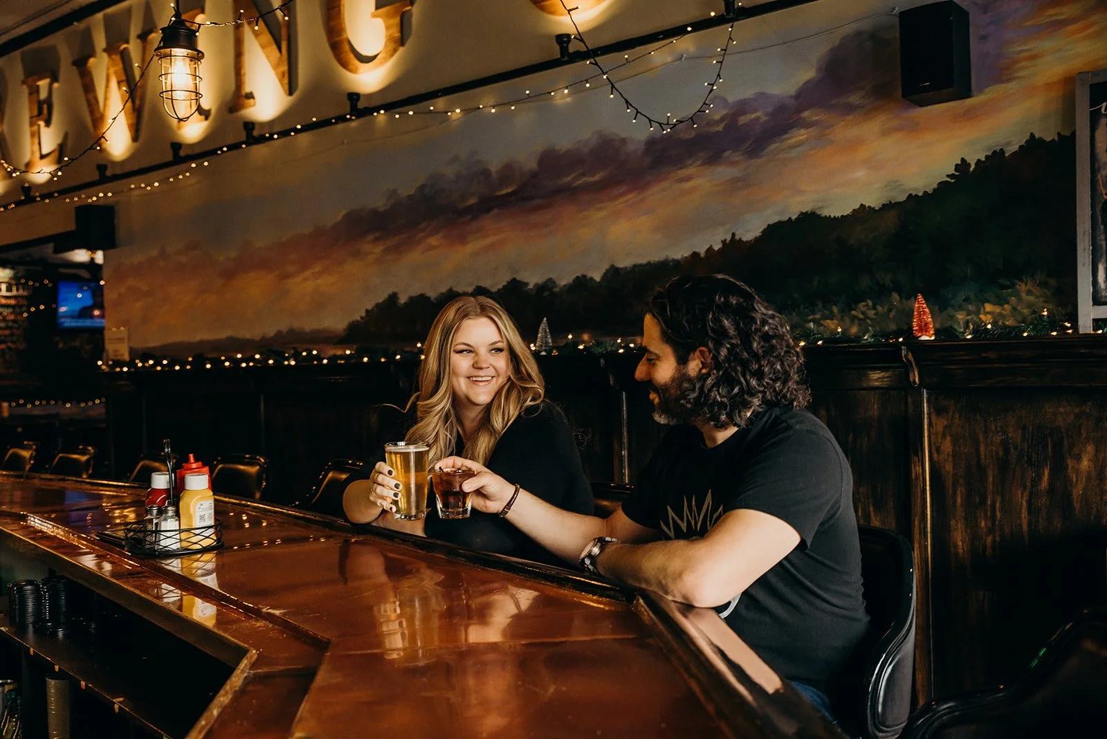A man and woman sitting at a bar in a restaurant, holding glasses of beer and a drink, smiling and enjoying their time together. The bar has condiments and napkins, with a painted mural of a sunset or sunrise on the wall behind them.