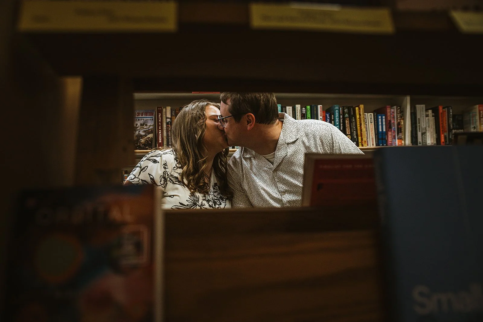 A couple kissing inside a bookstore, viewed through wooden shelves with books.