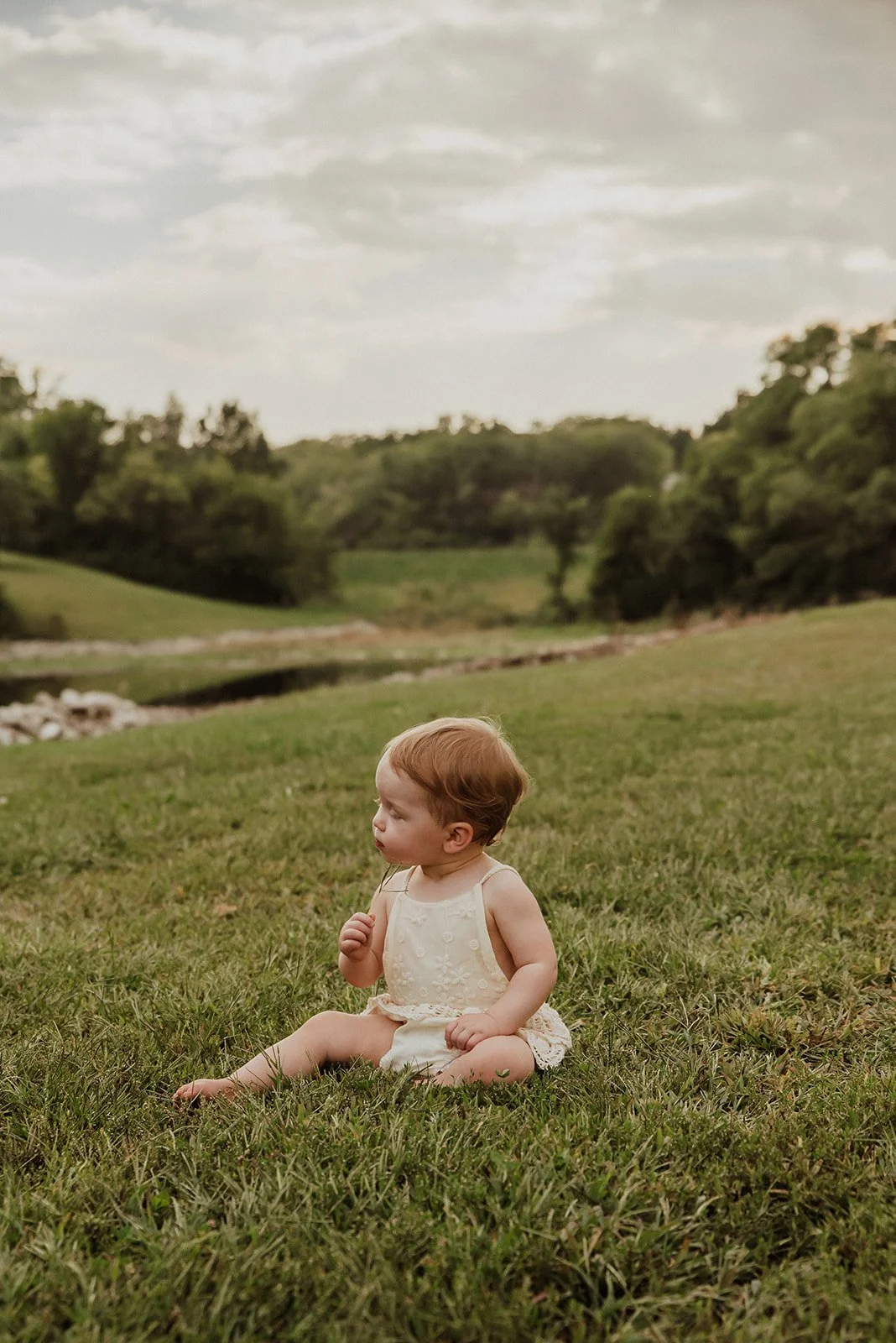 A young child with curly red hair and light skin sitting on green grass near a river in a countryside setting, with trees and hills in the background under a cloudy sky.