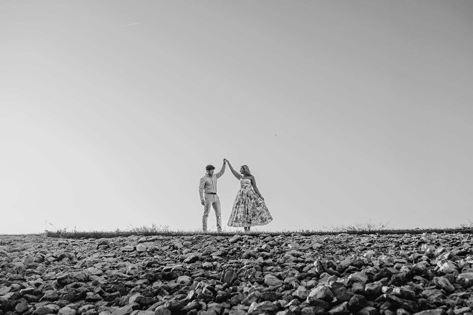A couple standing on a rocky ground holding hands in the air in a dance pose, with a clear sky background.