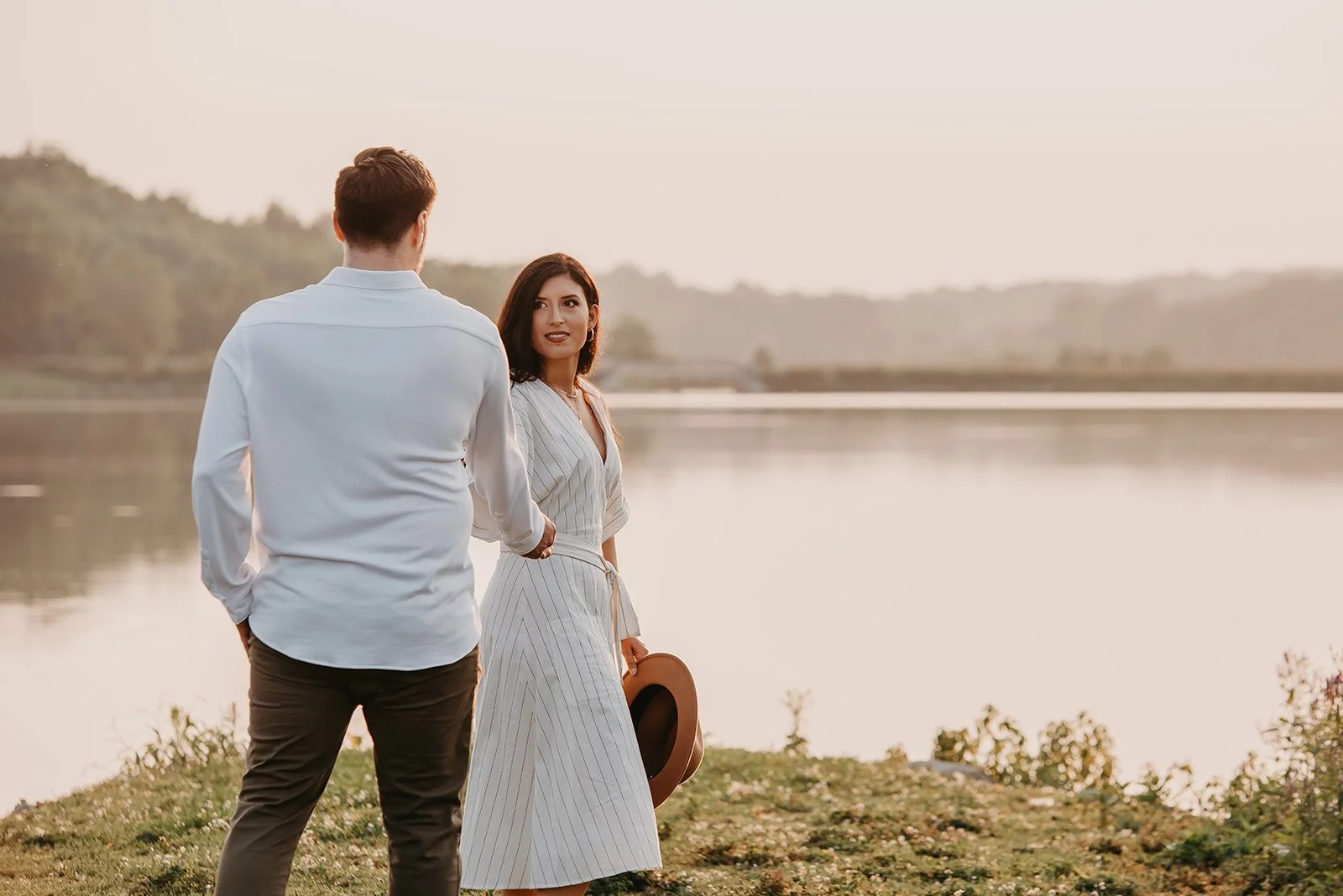 A man and woman standing by a lake during sunset, engaging in conversation. The woman holds a hat and looks at the man, who faces away from the camera. The scene is serene and picturesque.