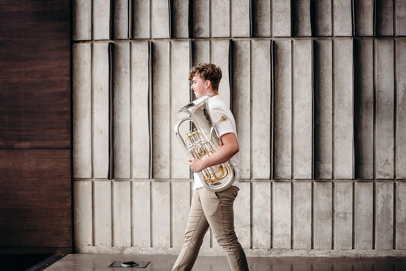 A young man with curly hair wearing a white shirt and khaki pants, standing sideways, holding a silver euphonium, against a textured concrete wall.