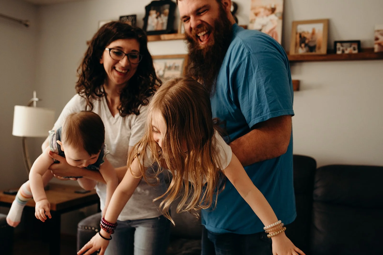 A family of four enjoying a playful moment in their living room. The mother is holding the young child while the father and daughter are bent over, laughing and engaging in the activity.