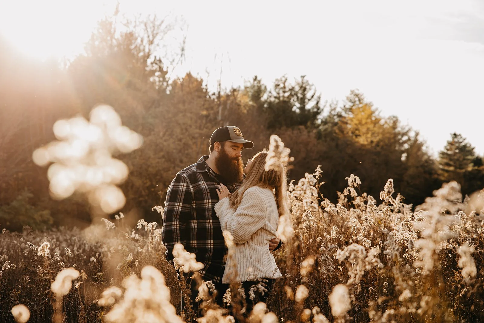A couple embraces in a field of tall, white flowers during golden hour, with trees in the background and sunlight creating a warm glow.