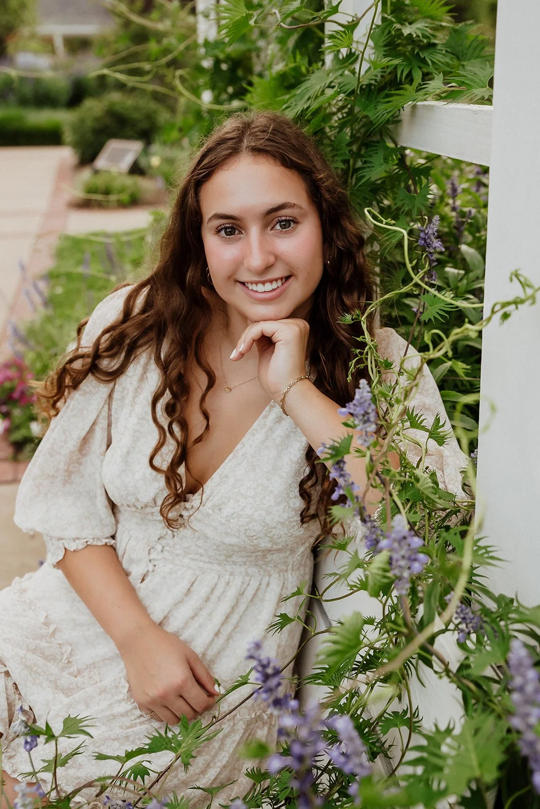 A young woman with long, curly brown hair smiling, resting her chin on her hand, in a garden with green plants and purple flowers.