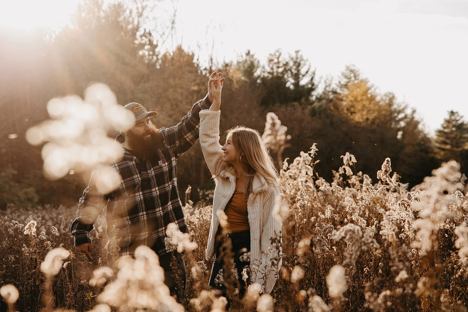 A man and a woman dancing hand in hand in a field of tall, brown wildflowers during sunset, with a forested background.
