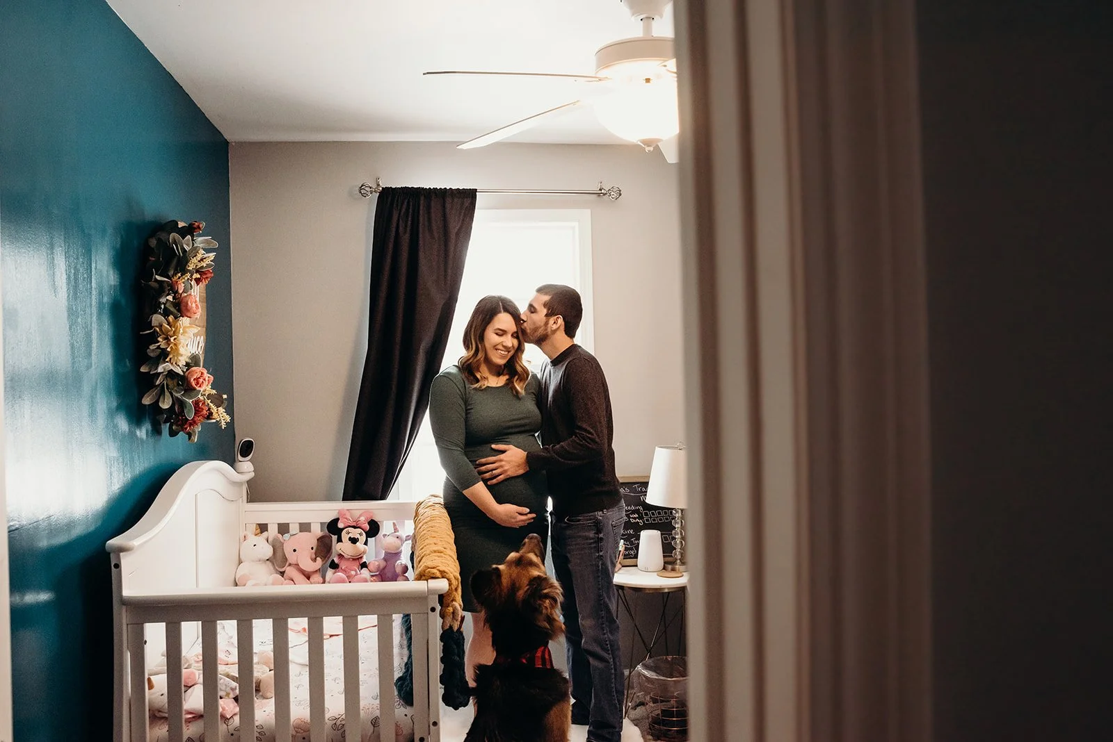 A couple standing in a nursery, the woman pregnant, with a dog looking up at them. The nursery has plush toys, a bed, and a window with curtains.