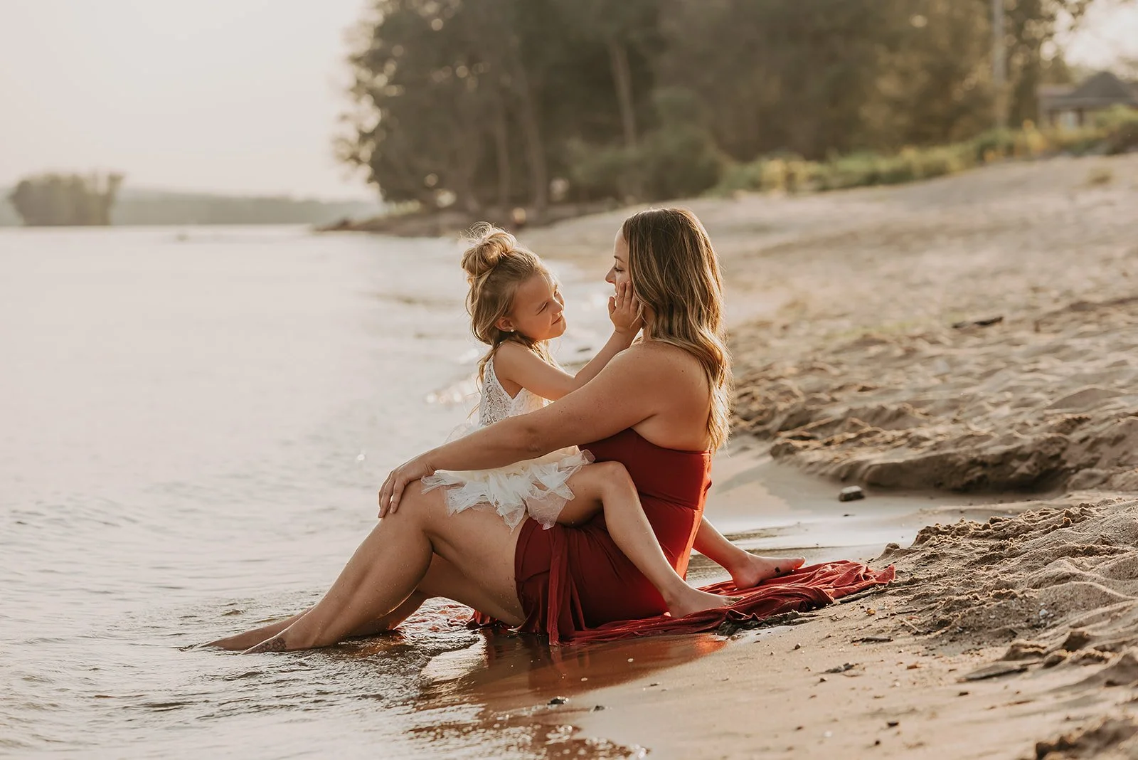 A woman and a young girl sitting on a sandy beach near the water, touching each other's faces, with trees and houses in the background during sunset.