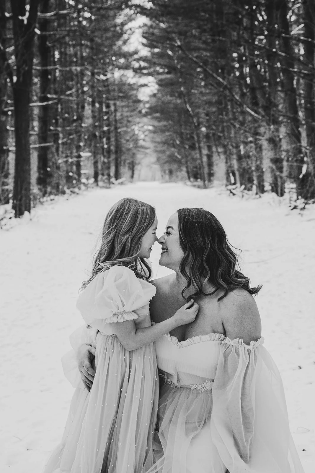 A black-and-white photo of a woman and a young girl touching noses in a snowy forest.