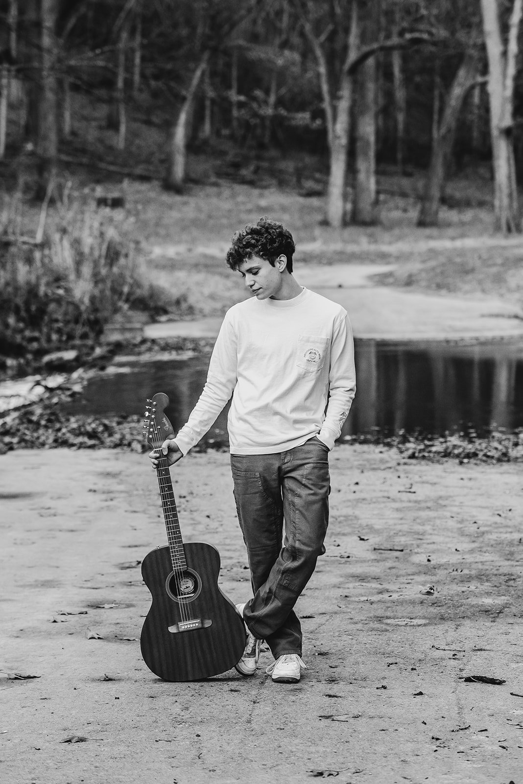 A young person with curly hair standing outdoors on a dirt path next to a guitar, looking down with a contemplative expression, surrounded by trees and a small body of water in the background.