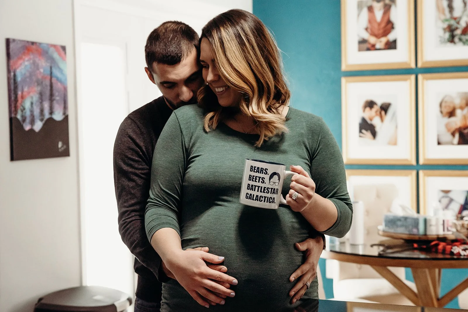 A couple embraces, with the man hugging the pregnant woman from behind. The woman is holding a coffee mug that says 'BEARS. BEETS. BATTLESTAR GALACTICA.' They are smiling and leaning their heads together, in a cozy indoor setting with framed photos o