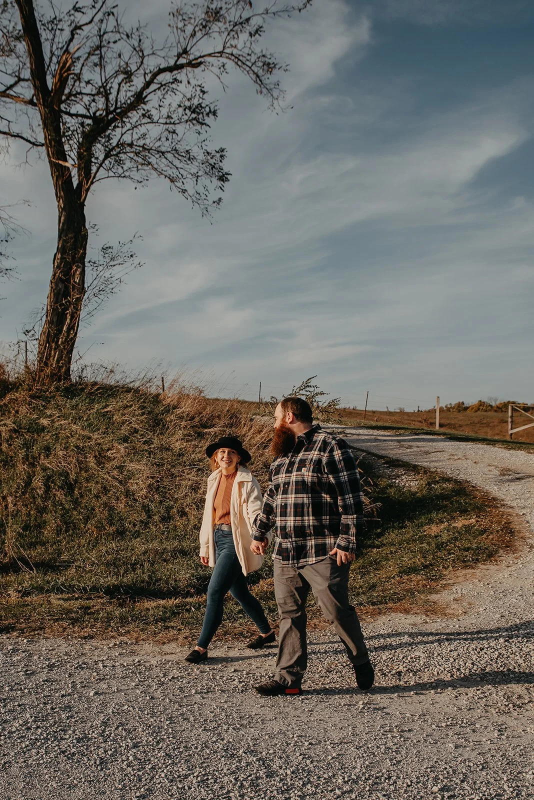 A smiling woman and a bearded man holding hands while walking on a gravel path in a rural outdoor setting during late afternoon or early evening, with a large bare tree on the left and open fields in the background.