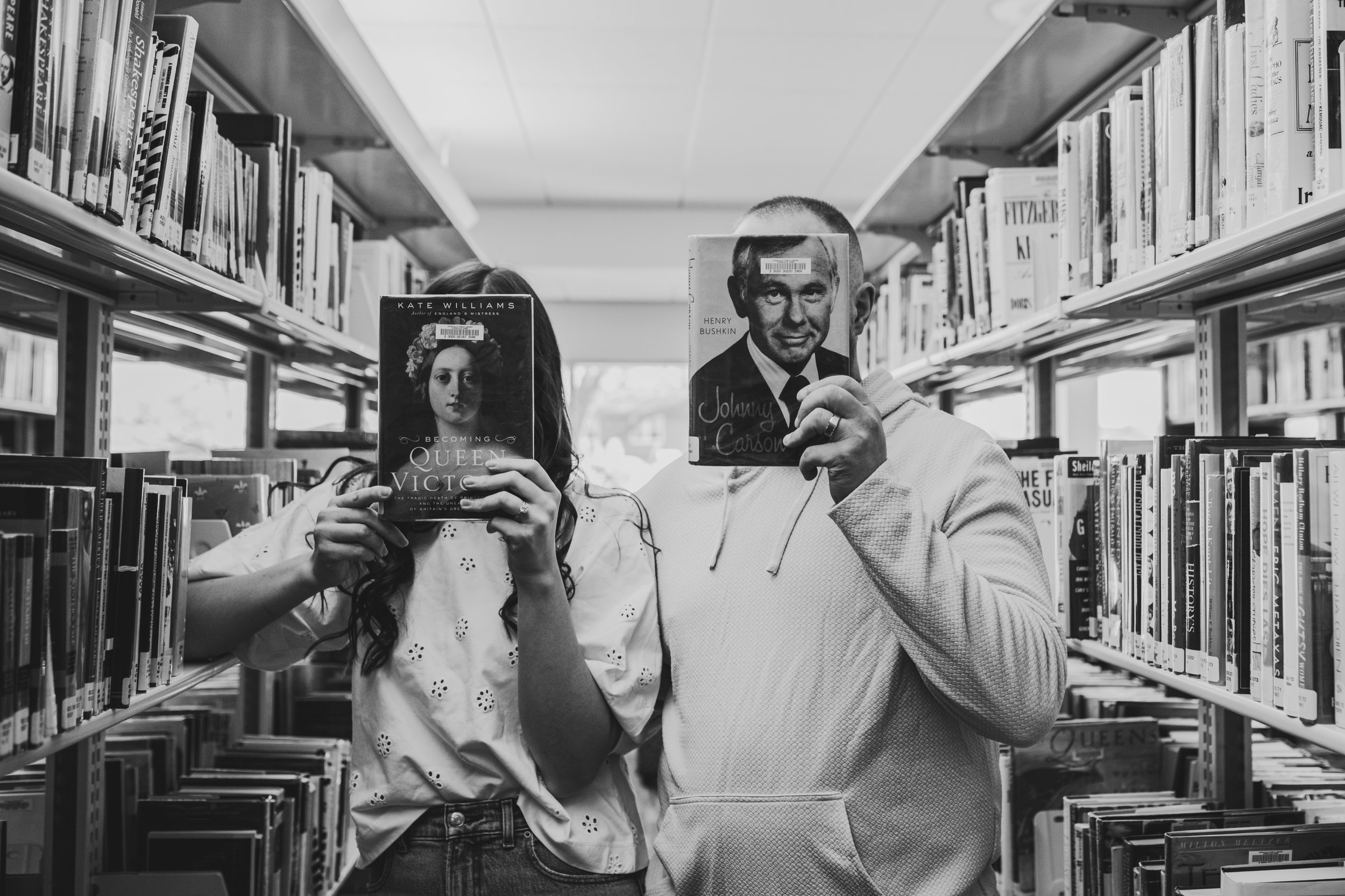 Two people standing in a library aisle, holding books in front of their faces. The woman on the left is holding 'Becoming Queen Victoria' by Kate Williams, and the man on the right is holding a book featuring Henry Bushkin's photo and the name Johnny