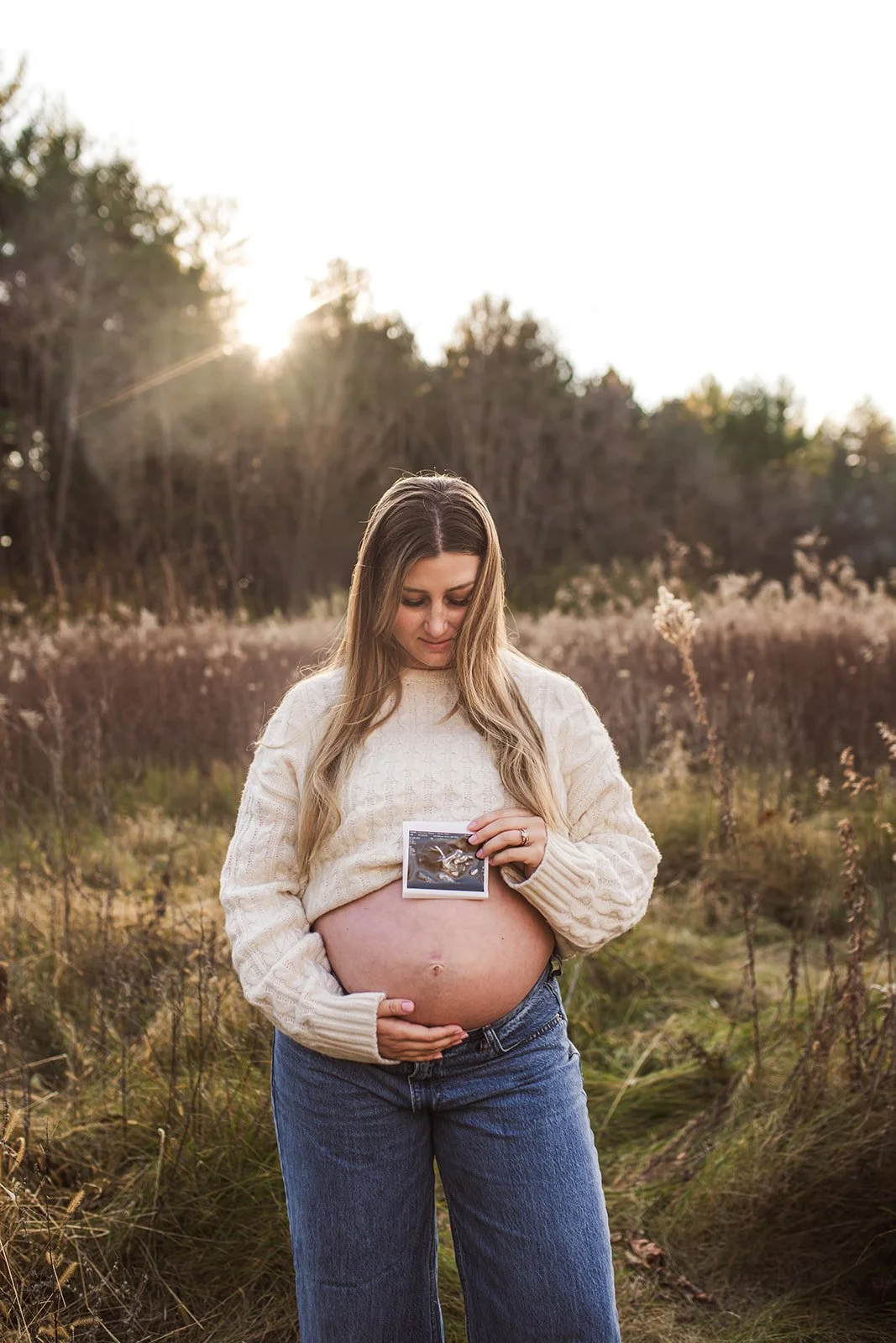 Pregnant woman outdoors holding ultrasound photo and showing her belly.
