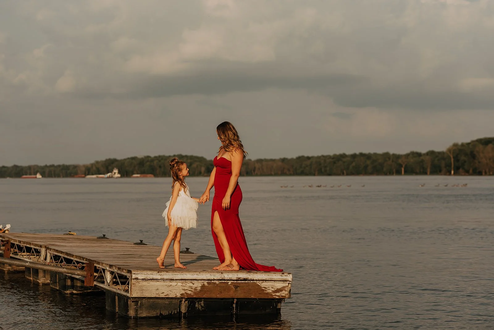 A woman and a young girl holding hands on a dock by the water, with boats and trees in the background, during sunset.