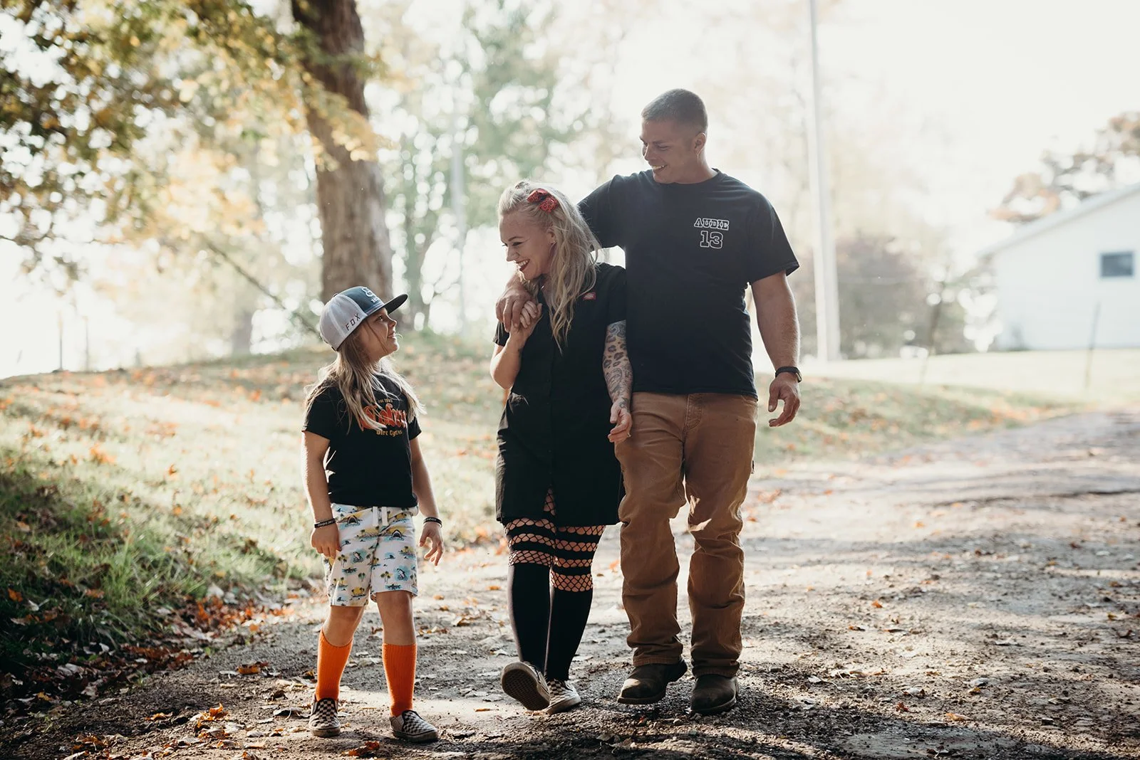 A family of three, including a woman, a man, and a young girl, walking on a dirt path in a park during autumn. They are smiling and holding hands, with the man and woman touching shoulders.