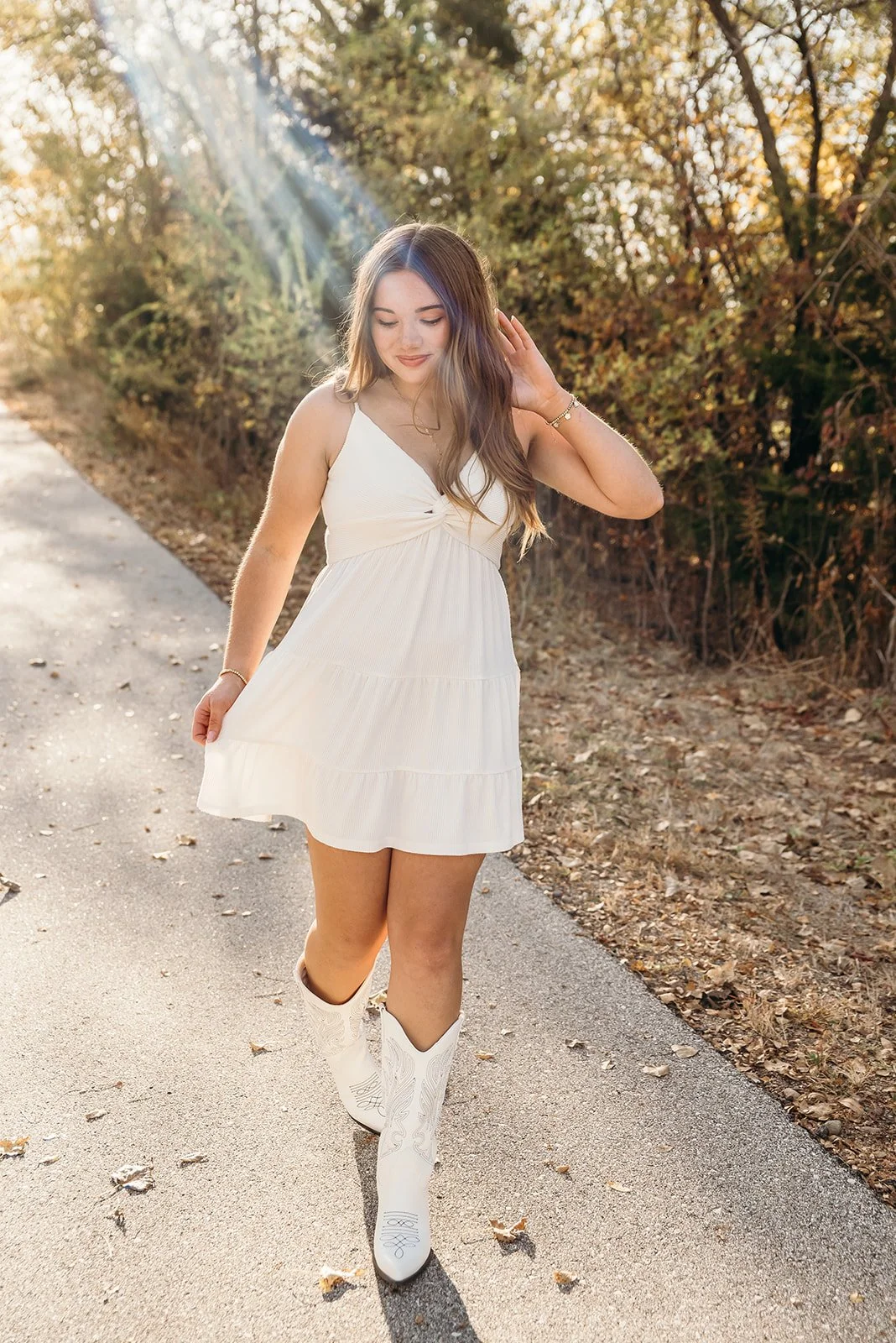 Young woman walking on a paved path outdoors in autumn, wearing a white dress and cowboy boots, with sunlight filtering through trees.