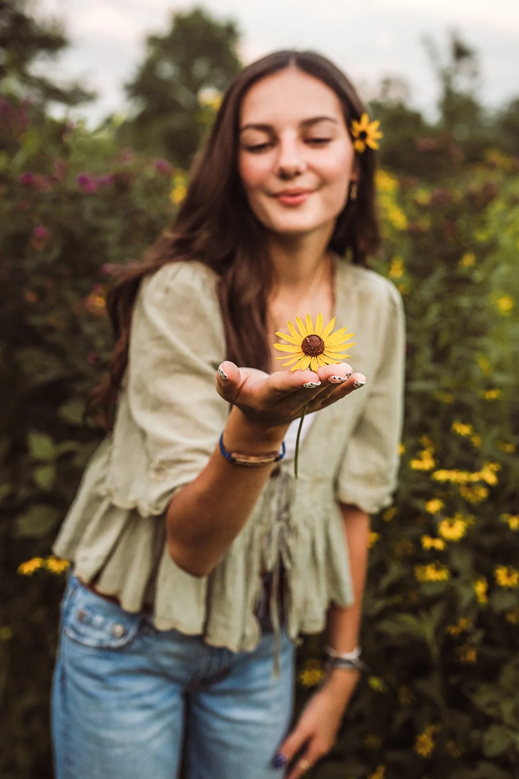 A young woman with long brown hair, holding a yellow flower in her hand, standing outdoors among yellow flowers, with a relaxed expression.