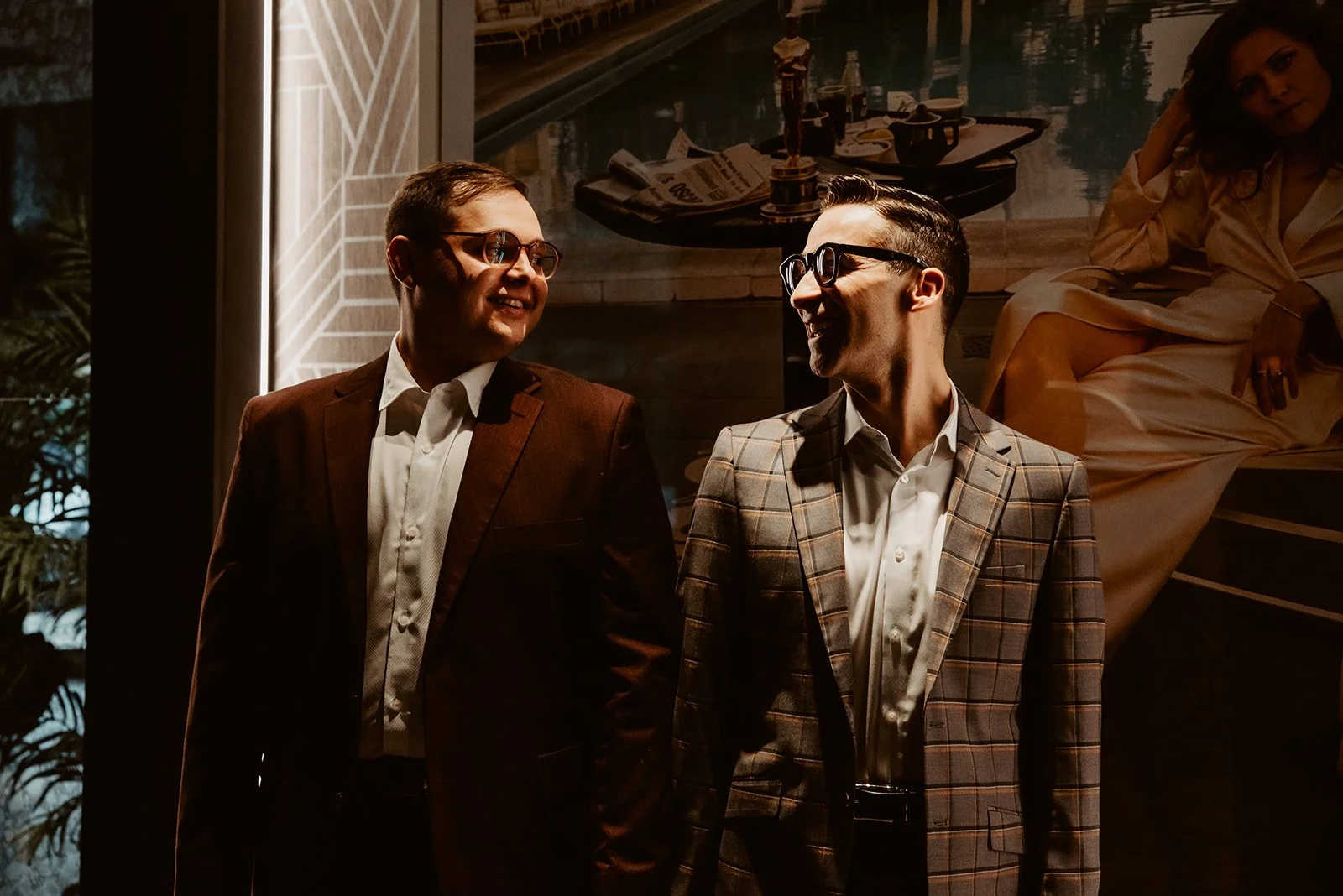 Two men in suits smiling and looking at each other, standing indoors with a woman sitting on a ledge in the background, and a painting of a boat and water on the wall behind them.