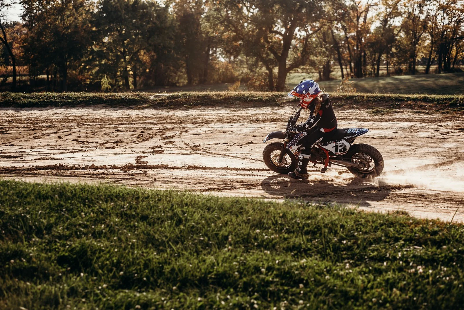 A person riding a dirt bike on a dirt track with trees and sunlight in the background.