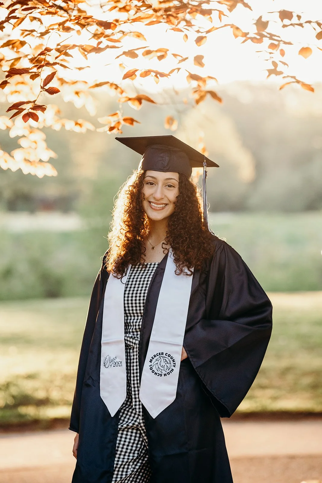 A young woman in a black graduation cap and gown, with a white stole, smiling outdoors during sunset, with trees and leaves in the background.