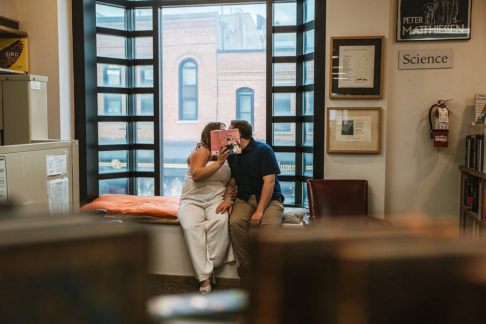 A couple sitting on a windowsill in a room, kissing while one person reads a pink book titled "Love Brain." The room has a large window, framed pictures, a fire extinguisher, and bookshelves.