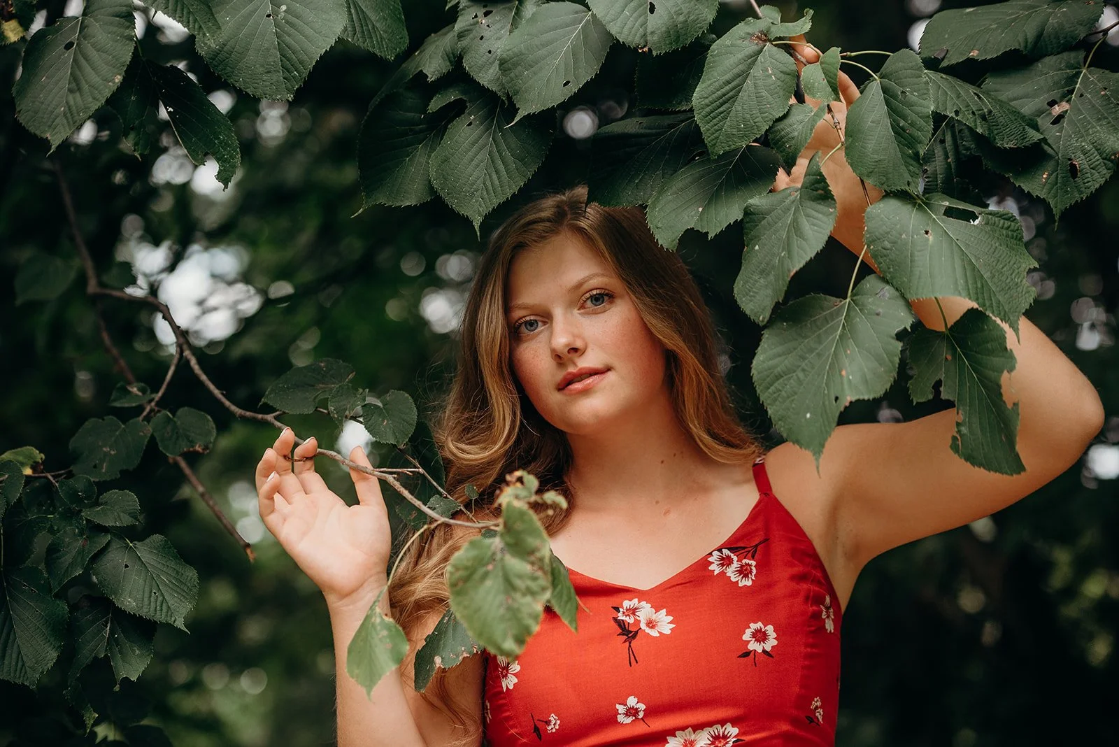 A woman with long wavy hair, wearing a red dress with white floral patterns, standing among green leaves and branches, with a blurred background of trees.