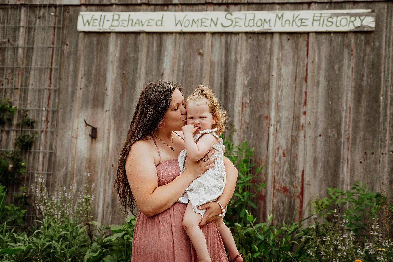 A woman with long brown hair wearing a pink dress is holding a young girl with curly blonde hair and a white dress. The woman is kissing the girl on the cheek. They are standing outdoors in front of a wooden fence with a sign that reads 'Well-Behaved