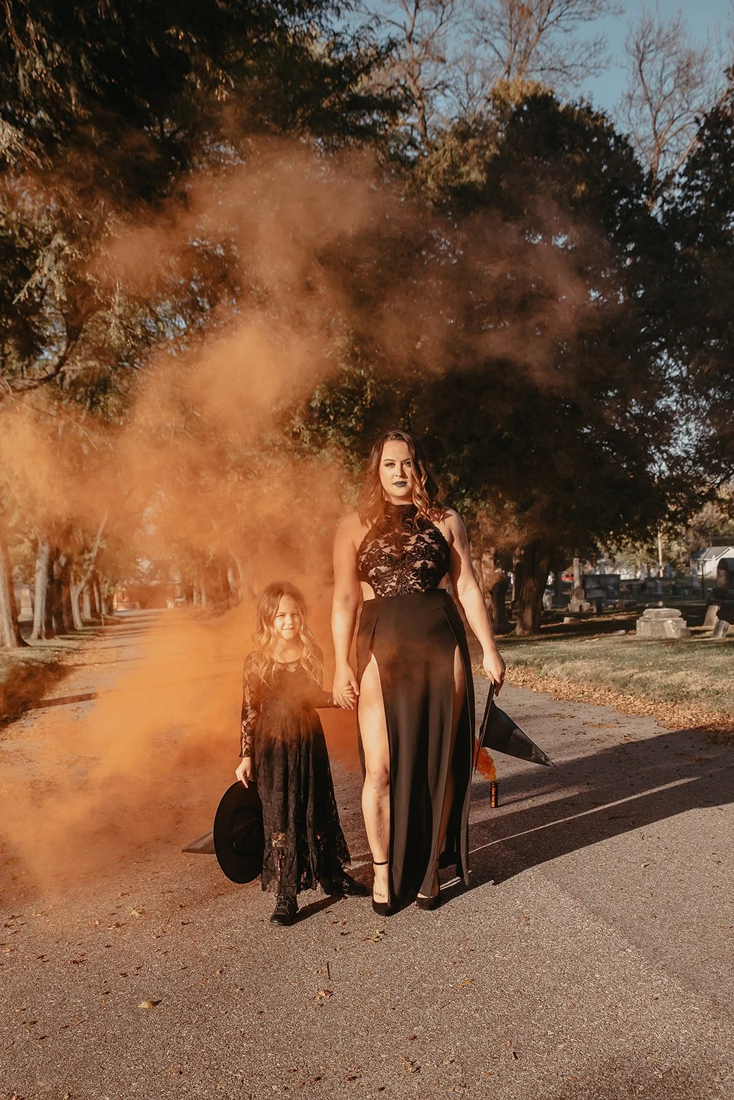 A woman and a young girl walking through a cemetery, holding hands, dressed in black lace dresses, with orange smoke in the background and trees lining the pathway.