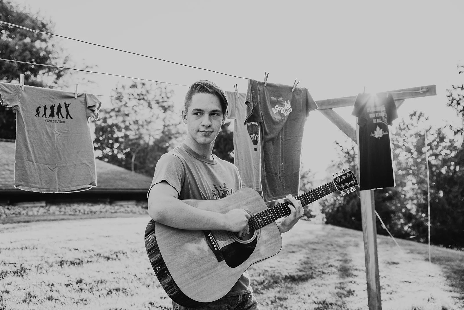 A young man with styled hair playing an acoustic guitar outdoors near a clothesline with shirts hanging on it, during a sunny day.