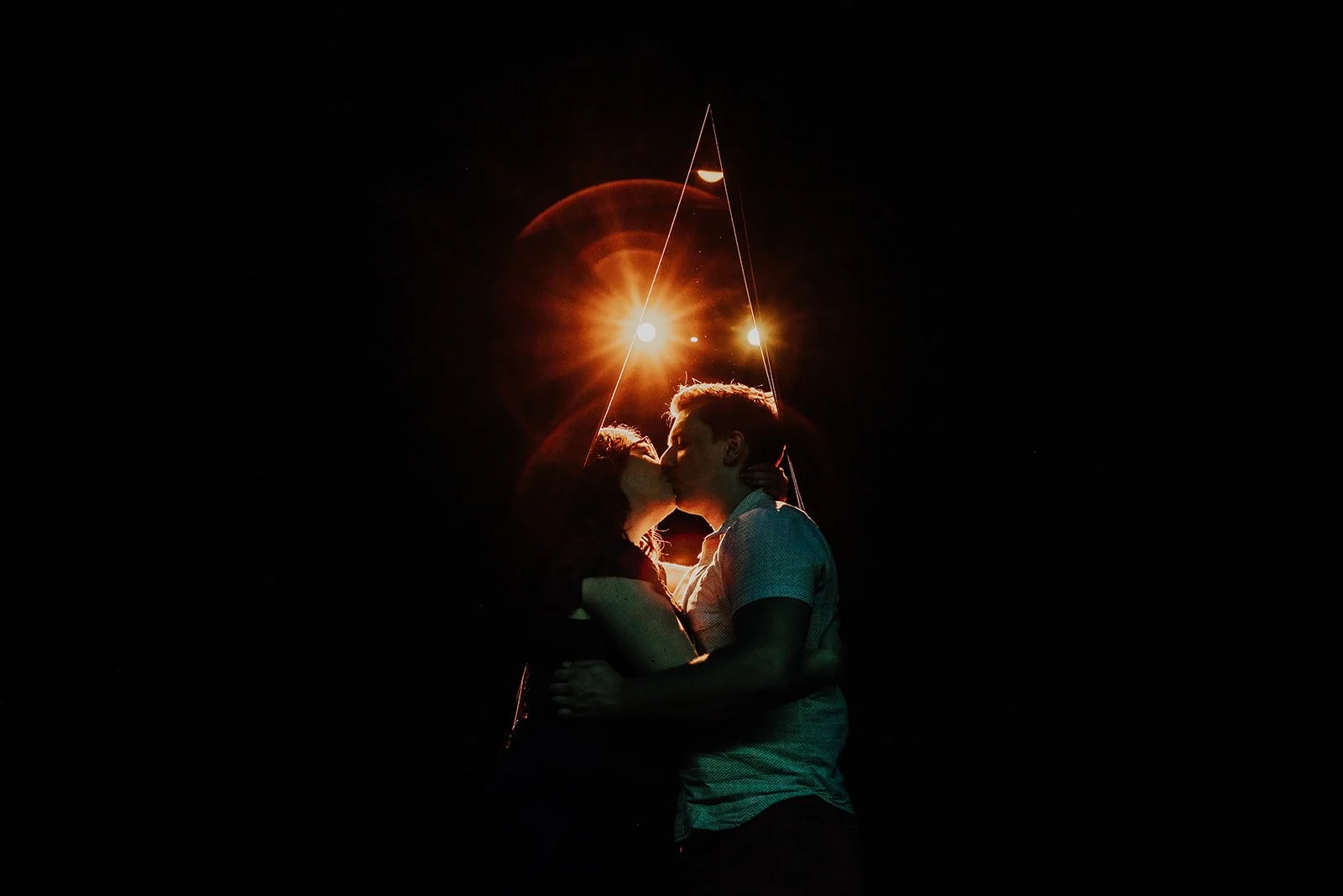 A couple kissing at night, backlit by bright lights and a streetlamp, with a dark sky surrounding them.