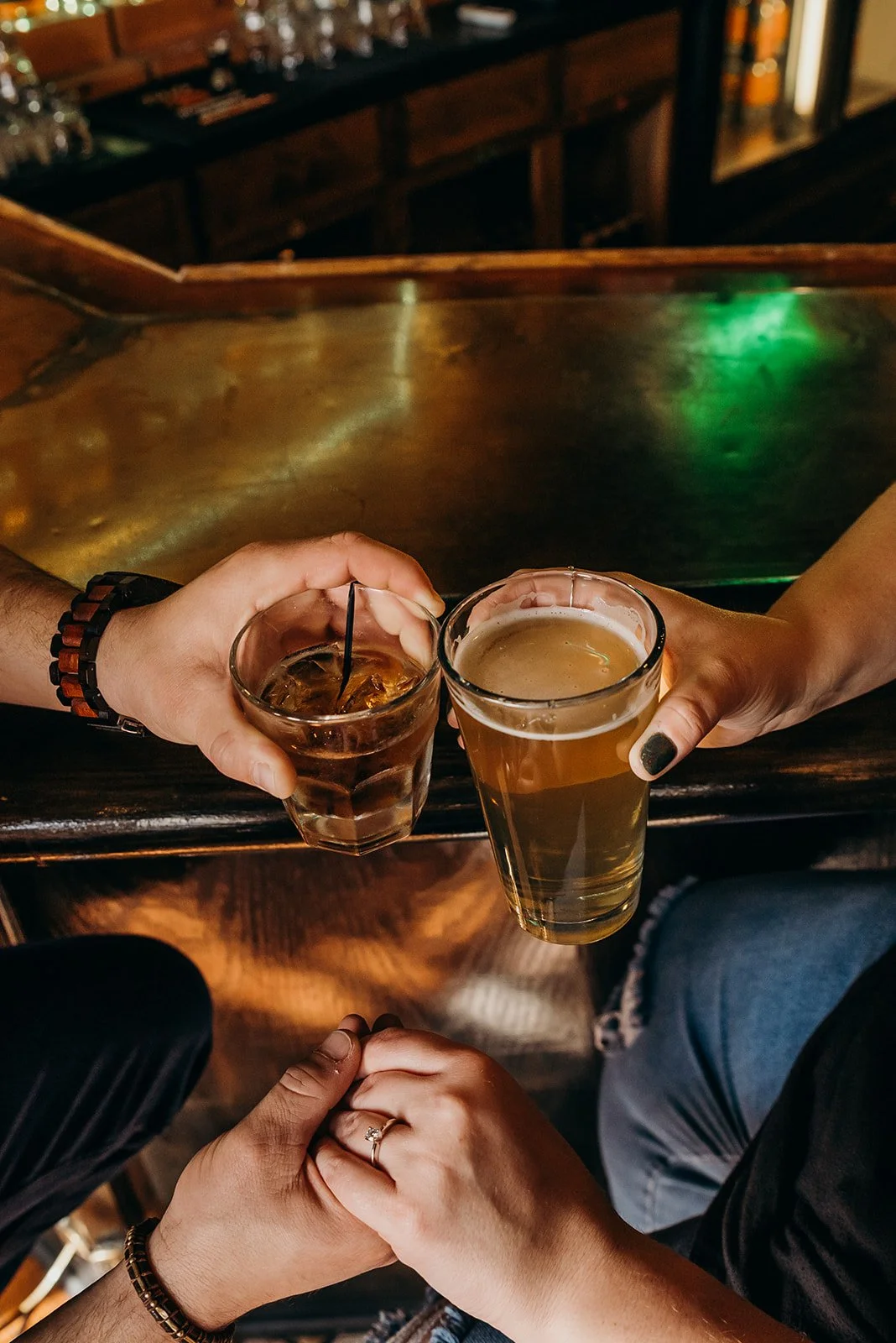 Two people holding drinks in a bar, one with a glass of whiskey and ice, the other with a glass of beer, with a bar counter in the background.