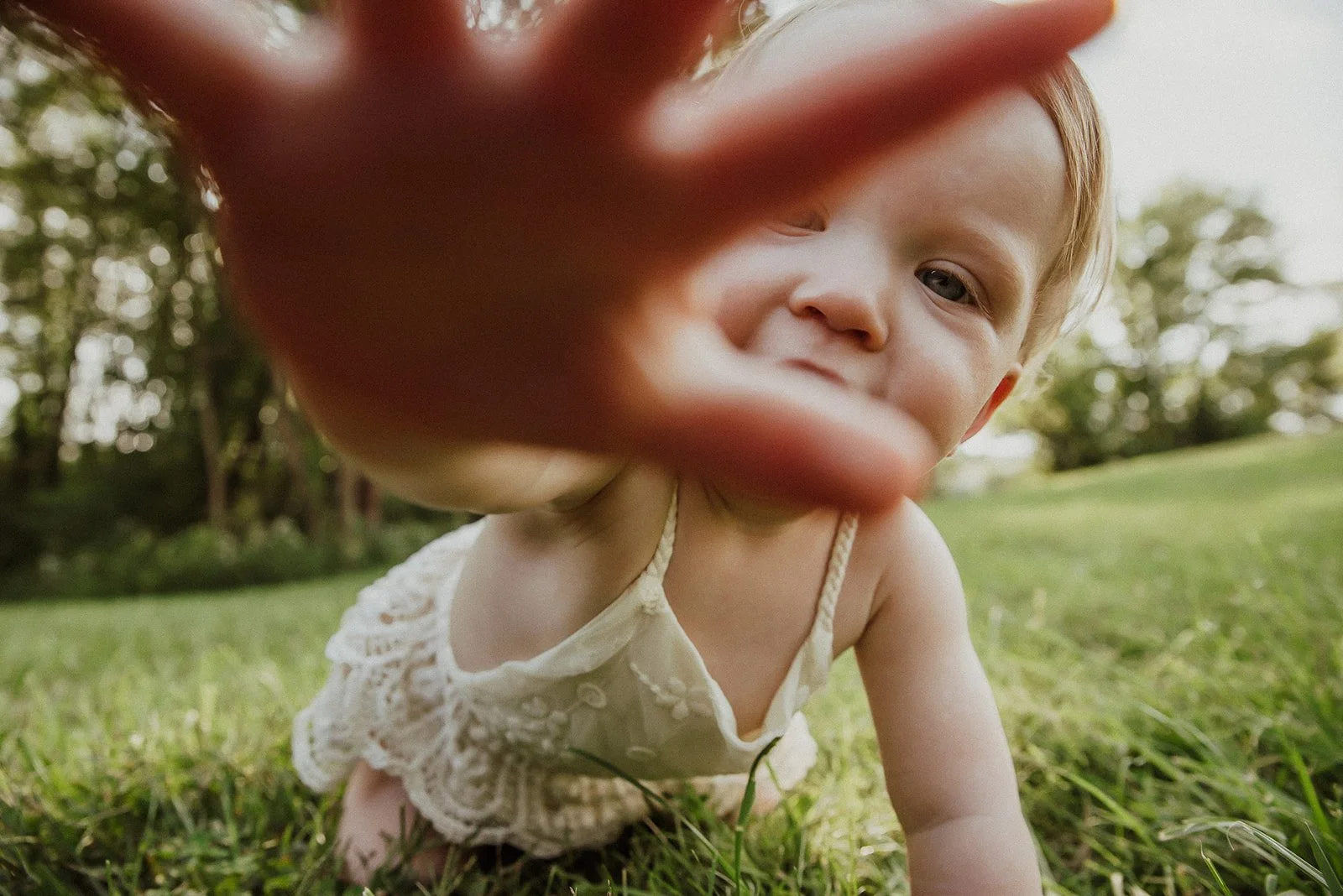 A baby crawling on grass outdoors reaching towards the camera, with a blurry background of trees and sky.