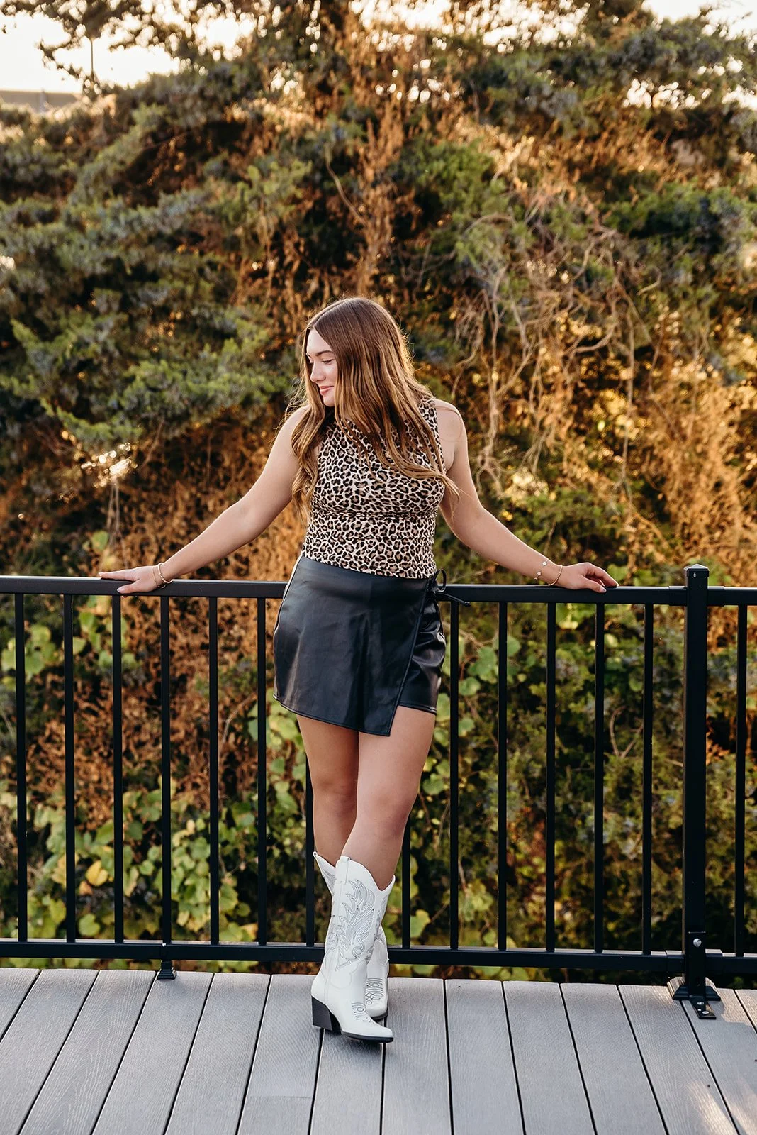 A young woman with long brown hair wearing a leopard print sleeveless top, a black leather skirt, and white cowboy boots standing on a balcony with a black railing, looking down and to the side, with trees and sunlight in the background.
