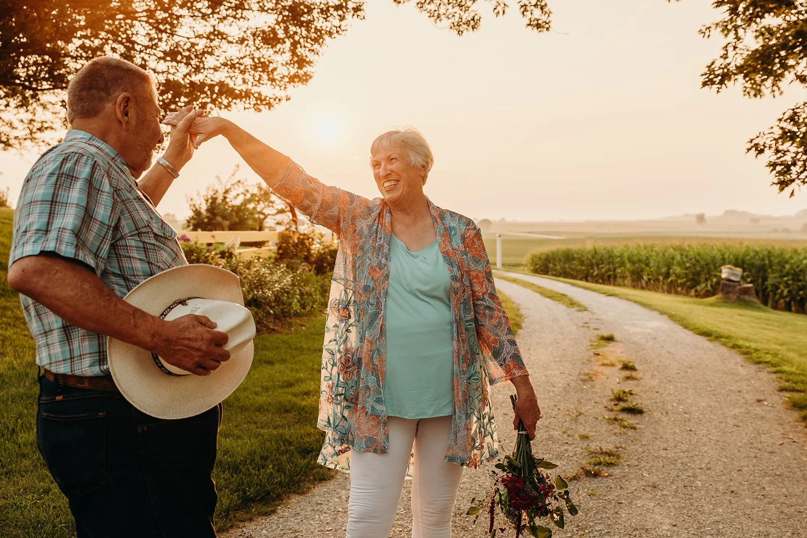 An elderly woman and man sharing a joyful moment outdoors at sunset, with the woman holding a bouquet of flowers and the man holding a hat, on a rural gravel path with fields and trees in the background.