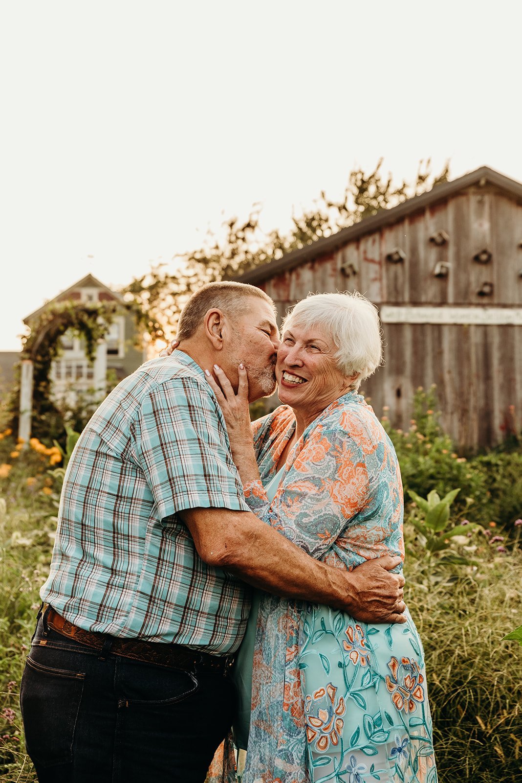 An elderly couple sharing a joyful moment outdoors in a garden with a rustic barn in the background, during sunset.