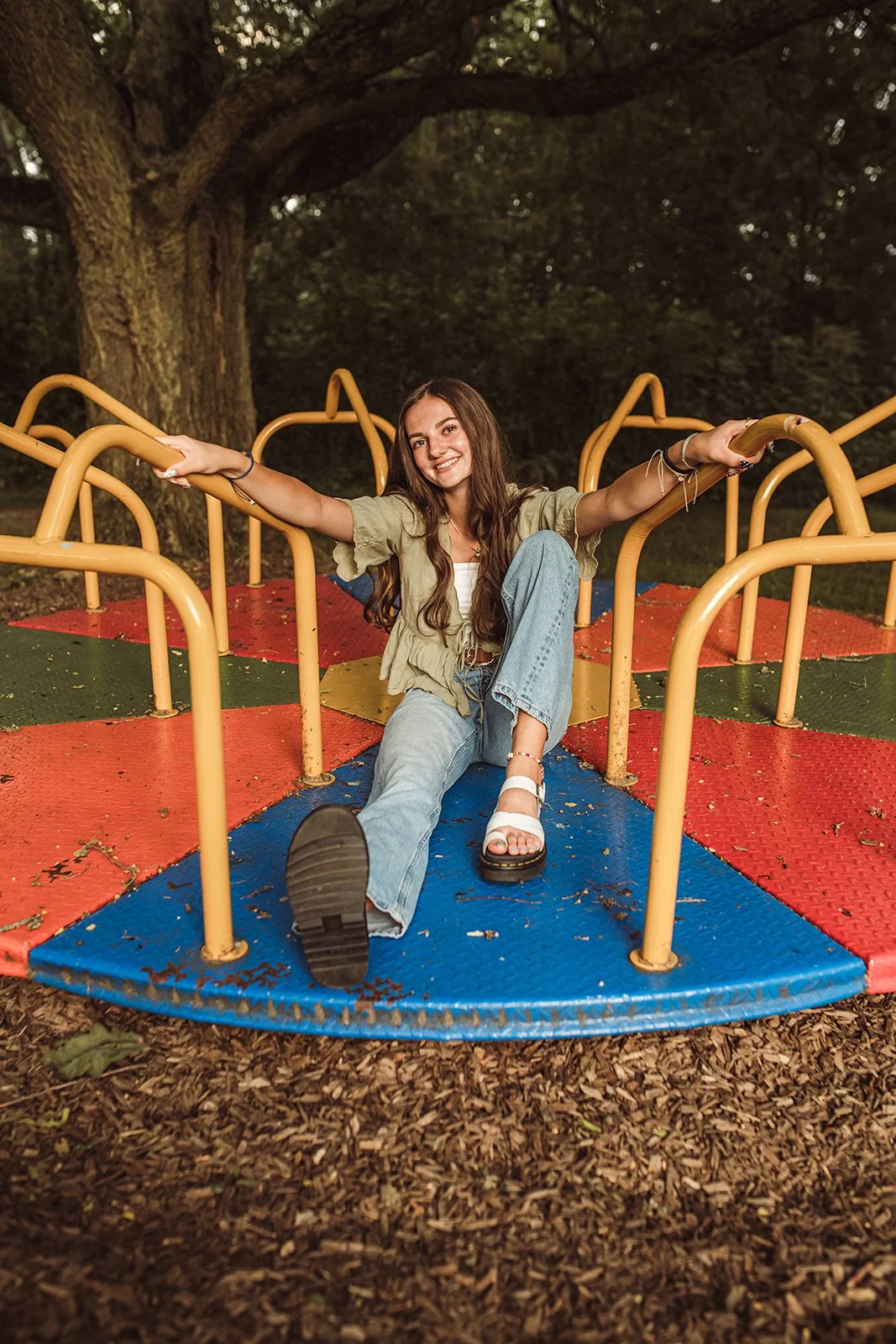 A young woman sitting on a colorful playground merry-go-round with yellow bars, smiling at the camera. She is wearing a beige jacket, white top, and blue jeans, with white sandals. The playground is set outdoors with a large tree in the background.