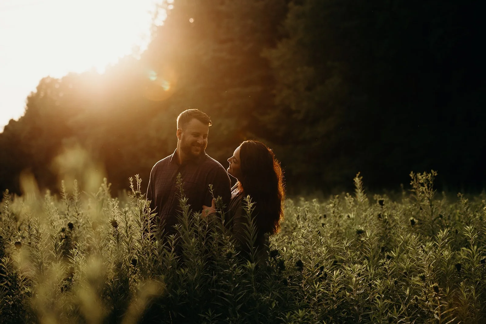 A smiling couple standing close together in a field of tall plants at sunset, with the sun setting behind them.