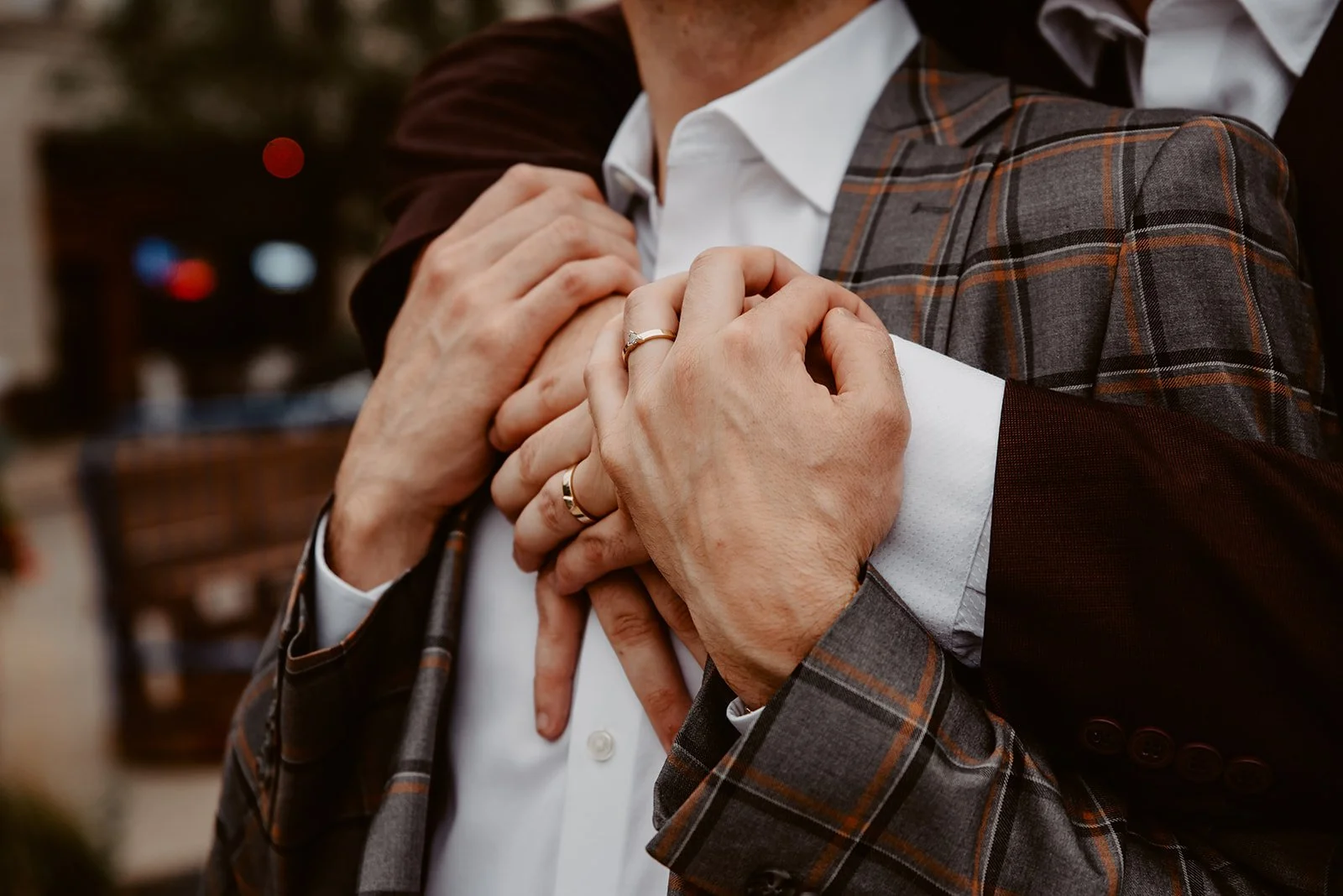 Two men in suits sharing a hug, with one man hugging the other's chest from behind. The man in front is wearing a white shirt and a gray plaid blazer, while the man behind is dressed in a dark suit.