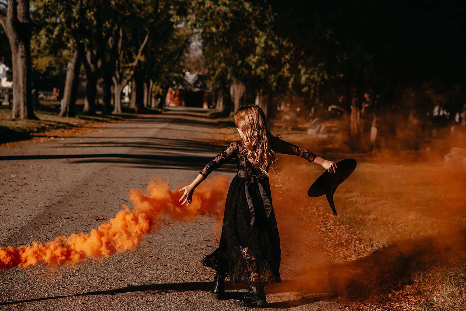 A girl in a black dress holding a black hat, releasing orange smoke on a street lined with trees.