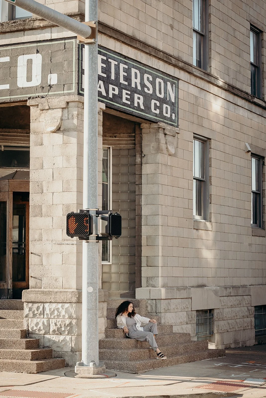 A woman with curly hair sitting on the steps outside a brick building with a sign that reads 'Petersen Paper Co.' near a street corner.