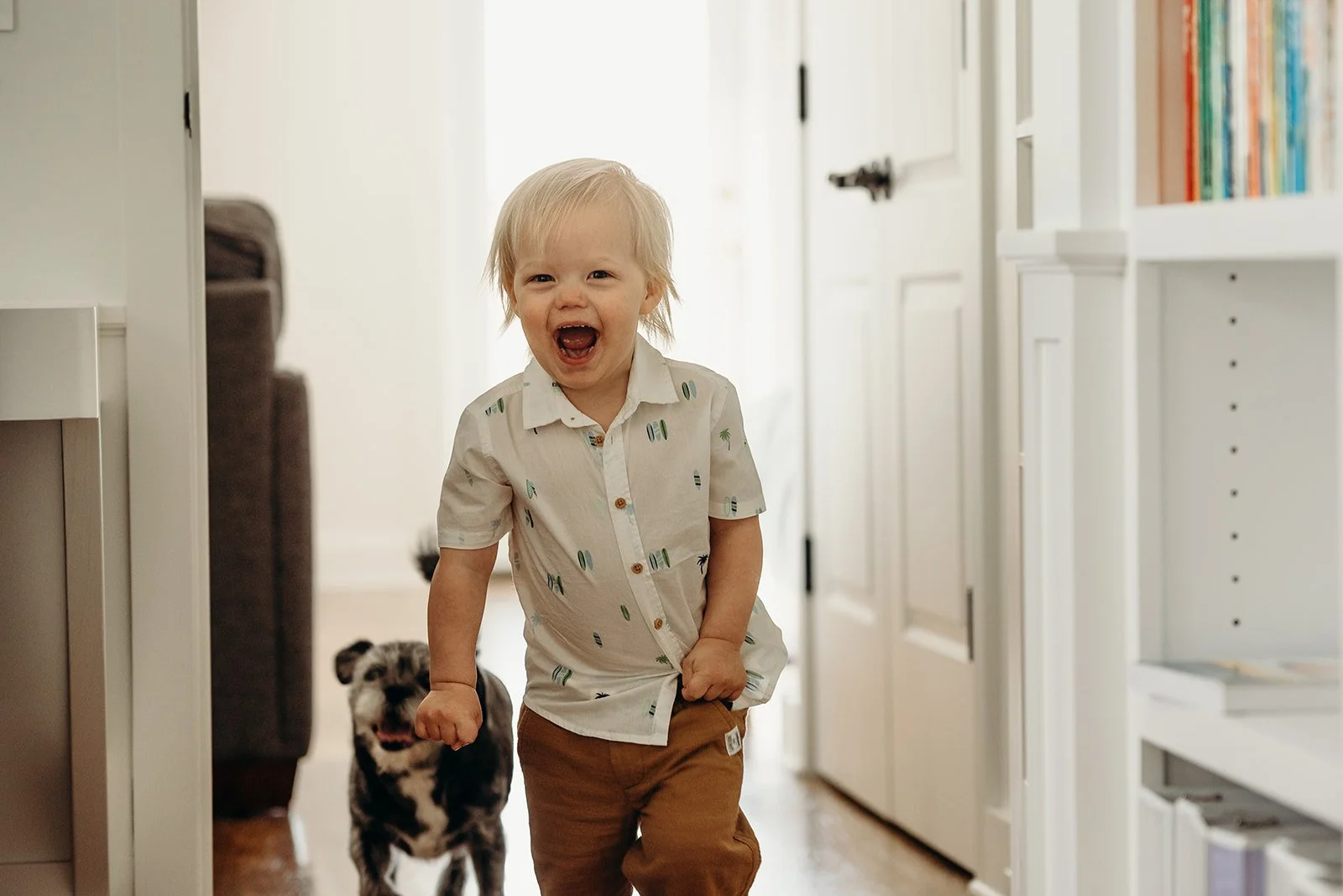 A young child with blonde hair, smiling and running in a hallway, with a small dog behind them. The child is wearing a white shirt with colorful pattern and brown pants.