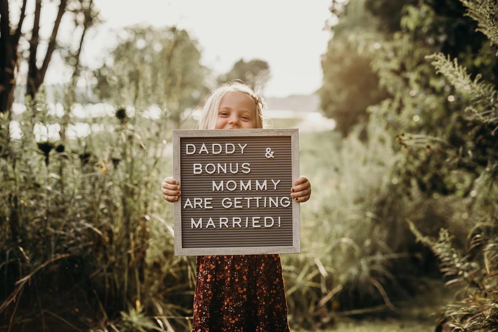 A young girl standing outdoors in a natural setting, smiling, holding a sign that says 'Daddy & Bonus Mommy Are Getting Married!'