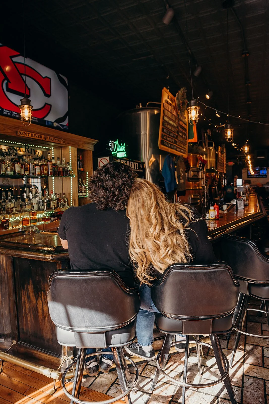 A couple sitting at a bar in a dimly lit pub, with their heads close together. The bar has various bottles of alcohol on display behind them and a wooden bar counter. The atmosphere is cozy and warm, with string lights and hanging lanterns.
